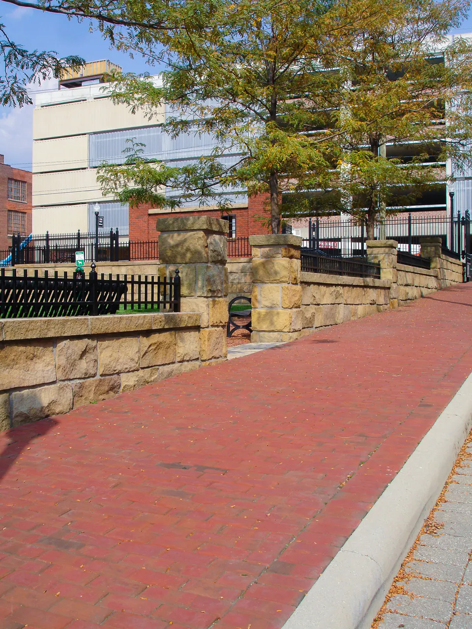 Brick walk beside stone walls, black railings, trees, and urban commercial buildings.